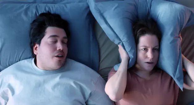 Two people lying in bed on blue pillows, with one person holding a pillow over the side of their head.