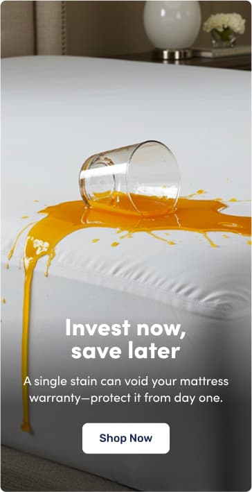 A glass of orange liquid has spilled onto a white mattress, showcasing the protective benefit of using a mattress protector. Text on the image reads: "Invest now, save later. A single stain can void your mattress warranty—protect it from day one." A button at the bottom says "Shop Now."