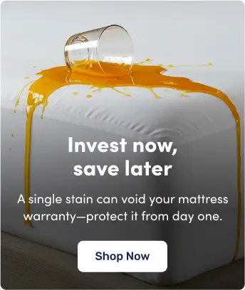 A glass of orange liquid has spilled onto a white mattress, showcasing the protective benefit of using a mattress protector. Text on the image reads: "Invest now, save later. A single stain can void your mattress warranty—protect it from day one." A button at the bottom says "Shop Now."