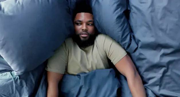 Person resting in bed with their head between blue pillows and covered by blue bedding.
